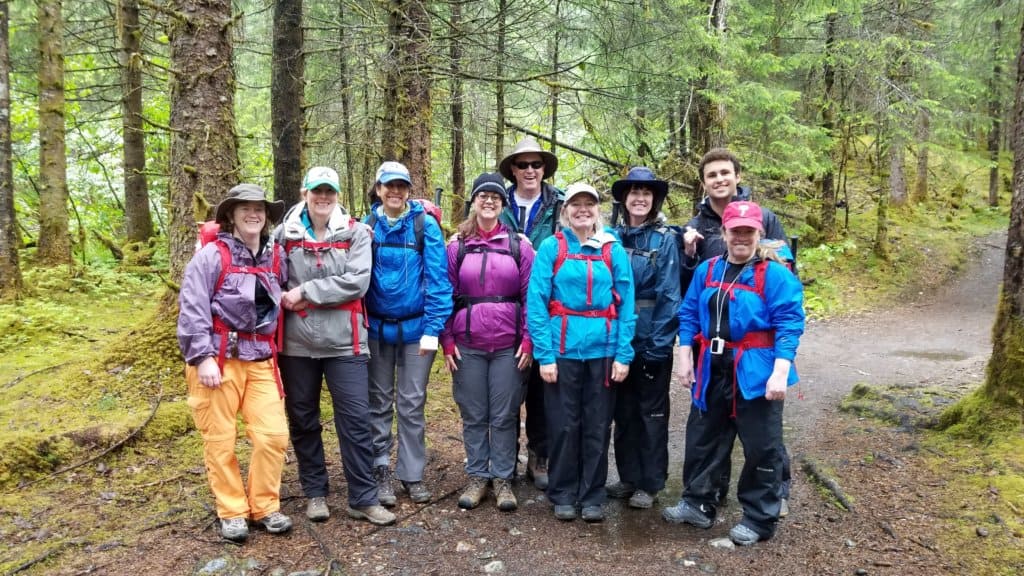 7 females and 2 males standing on a path in the woods all wearing rain jackets and backpacks