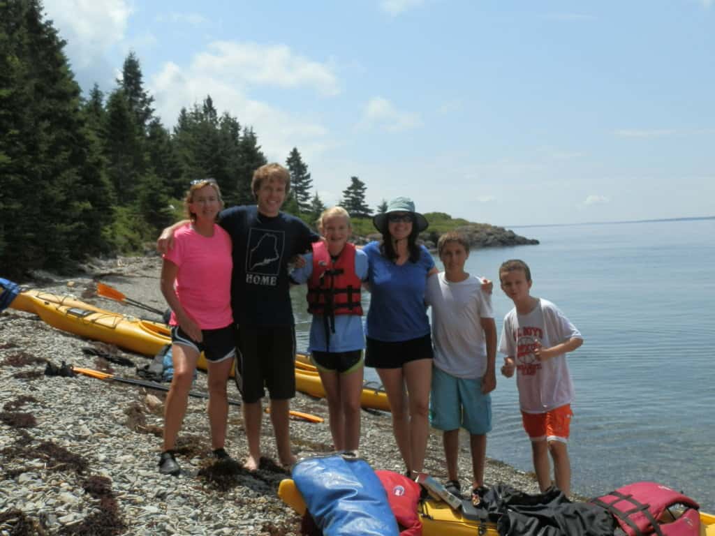 2 adults and 3 kids standing along side the water with kayaks in the background