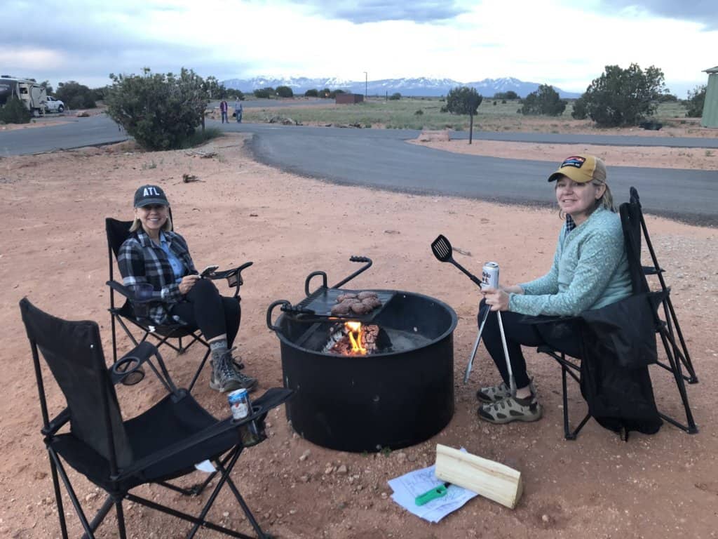 2 females sitting in camping chairs by a campfire cooking hamburgers